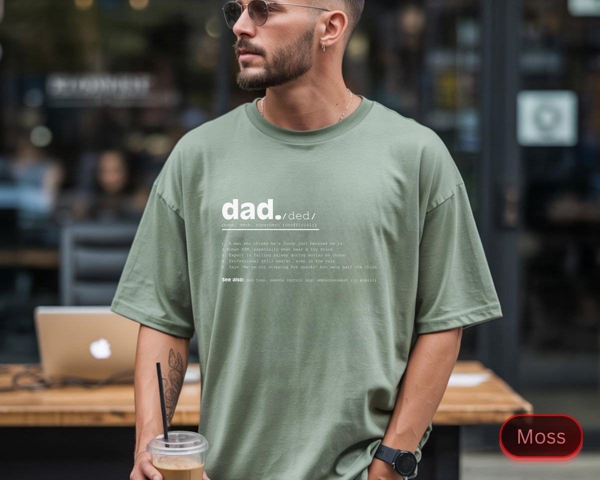 May include: A moss green t-shirt with the word "dad" and a definition printed in white. The shirt has short sleeves and a relaxed fit. The text is centered on the front of the shirt.