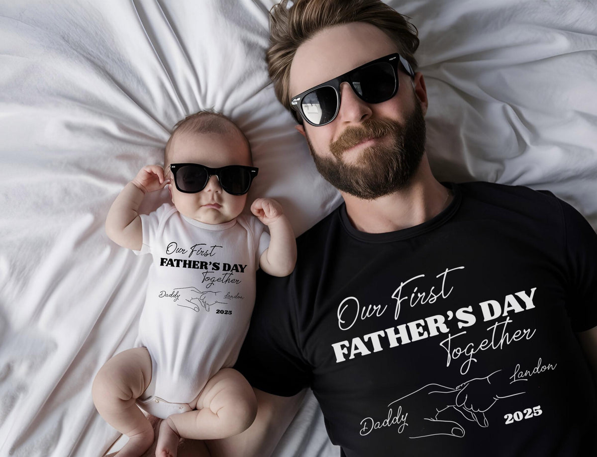 May include: A black t-shirt and a white baby onesie, both with the text "Our First Father's Day Together". The onesie and t-shirt also have a graphic of two hands holding each other. Both are wearing black sunglasses.