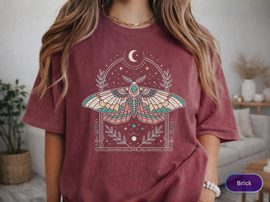 May include: Brick-colored t-shirt featuring a colorful moth design within a decorative frame. The moth has detailed wings with shades of teal, pink, and yellow. The design includes a crescent moon, stars, and botanical elements.