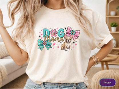 May include: Ivory-colored t-shirt featuring a colorful graphic. The design includes the text "Dog Mama" with a paw print, a French Bulldog illustration, and decorative bows. Pink and turquoise accents complete the design.