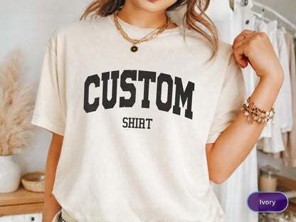 May include: Ivory-colored t-shirt with the word "CUSTOM" arched across the chest in bold, black letters, with "SHIRT" printed below. The shirt is short-sleeved and appears to be made of a soft material. The image also shows a gold necklace and bracelets.