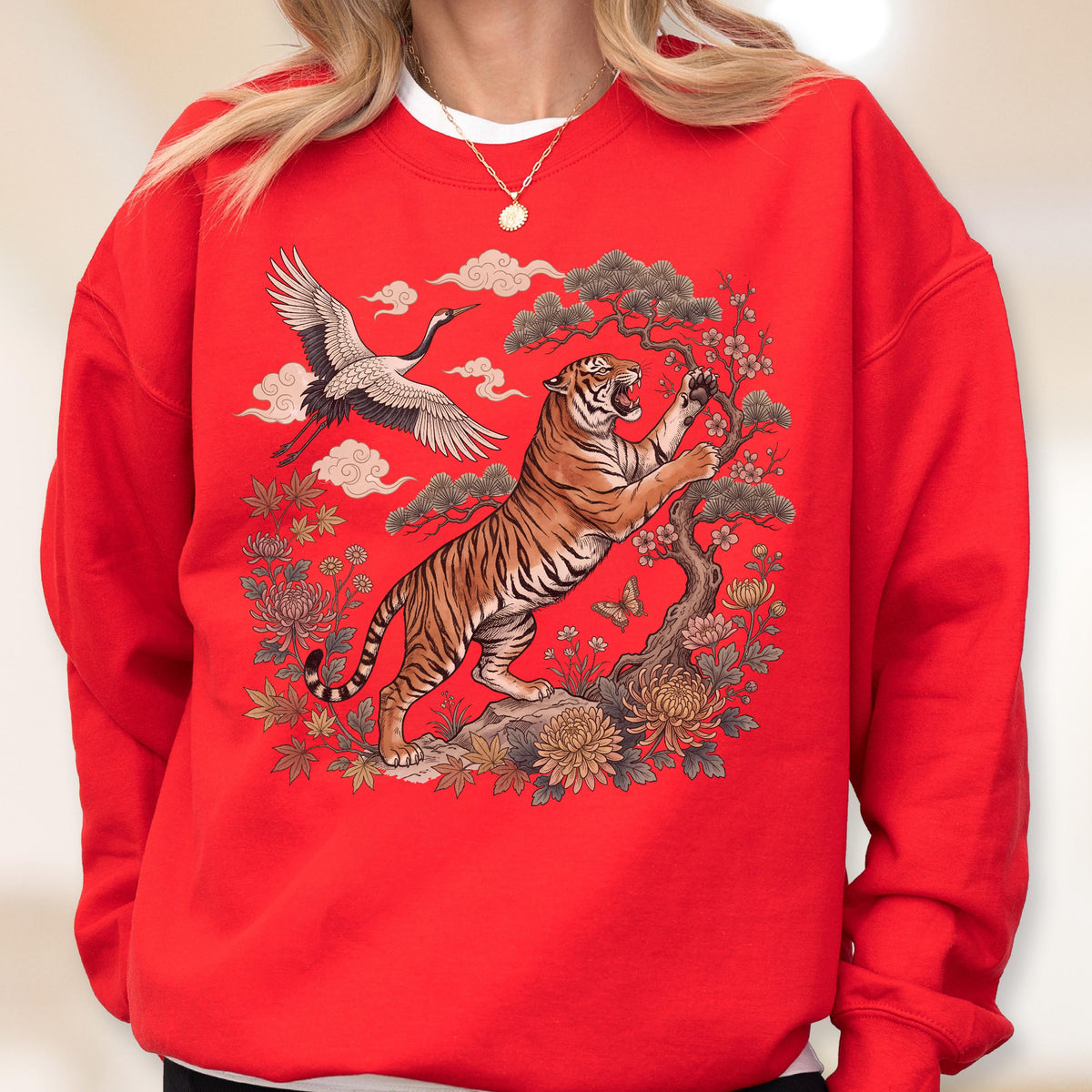 May include: Red sweatshirt featuring a detailed illustration of a tiger, crane, and floral elements. The tiger is depicted with orange and black stripes, reaching towards a tree. A crane flies in the background, with clouds and flowers completing the design.