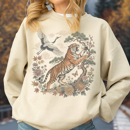 May include: Beige sweatshirt featuring a detailed illustration of a tiger climbing a tree, a crane in flight, and floral elements. The design incorporates a blend of orange, brown, and cream colors, with a touch of pink and green. The overall theme is inspired by Asian art.