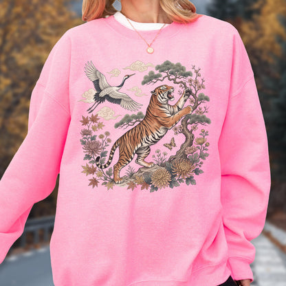 May include: Pink sweatshirt featuring a detailed illustration of a tiger, crane, and floral elements. The tiger is depicted with orange and black stripes, climbing a tree. A crane is flying above. The design is set against a pink background.