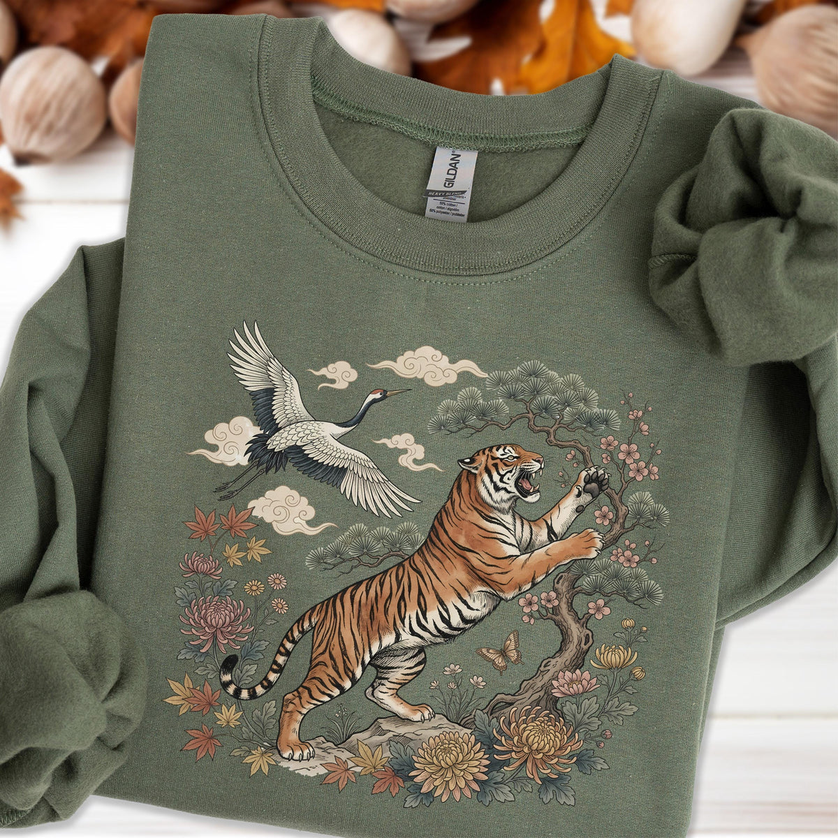 May include: An olive green sweatshirt featuring a detailed graphic of a tiger, crane, and floral elements. The tiger is orange and black, reaching towards a flowering tree. A white crane flies above, with clouds in the background.