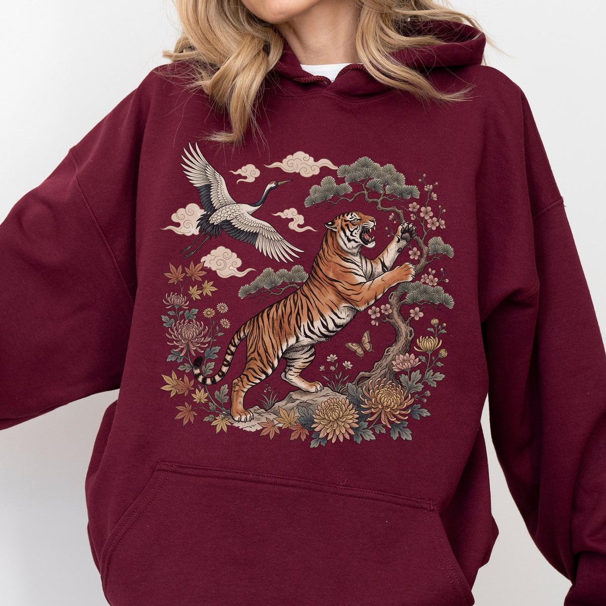 May include: Burgundy hoodie featuring a detailed illustration of a tiger, crane, and floral elements. The tiger is orange and black, set against a backdrop of clouds and flowers. The design has an Asian-inspired aesthetic, perfect for casual wear.