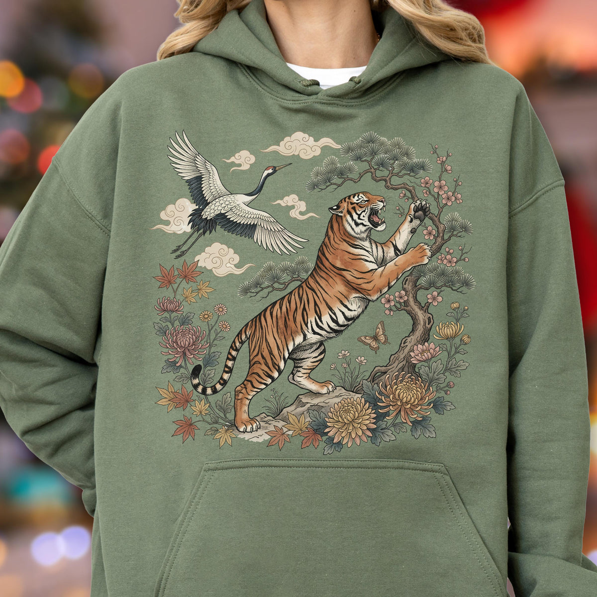 May include: Sage green hoodie featuring a detailed illustration of a tiger, crane, and floral elements. The tiger is depicted with orange and black stripes, reaching towards a tree. The design includes clouds and flowers, creating a nature-inspired theme.