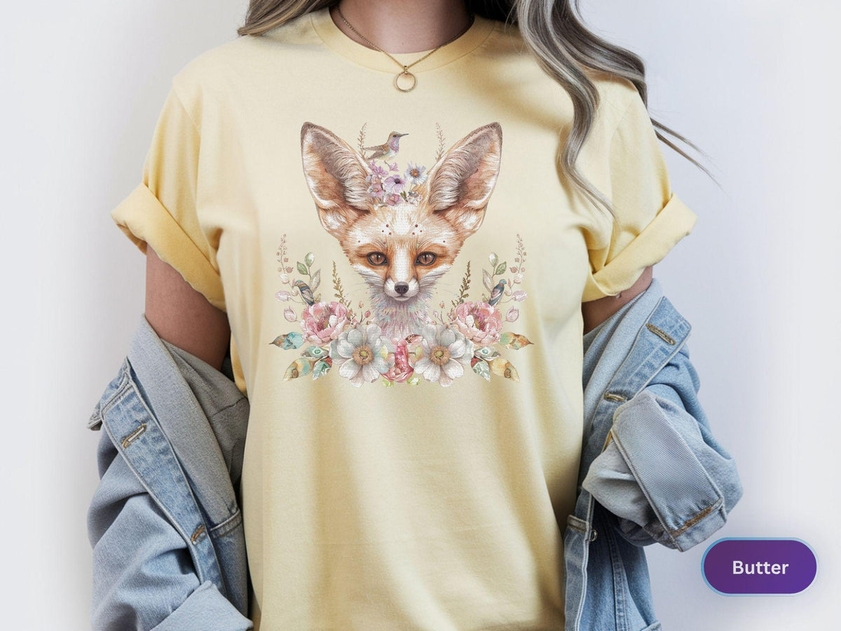 May include: Butter-colored t-shirt featuring a watercolor illustration of a fox with large ears, adorned with flowers and a small bird. The fox is the focal point, surrounded by floral elements in soft pastel colors. The shirt has rolled-up sleeves.