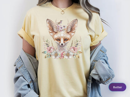 May include: Butter-colored t-shirt featuring a watercolor illustration of a fox with large ears, adorned with flowers and a small bird. The fox is the focal point, surrounded by floral elements in soft pastel colors. The shirt has rolled-up sleeves.