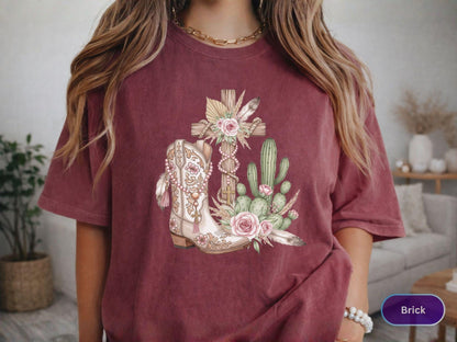 May include: Brick-colored t-shirt featuring a detailed graphic of a cowboy boot, a cross, and cacti. The boot is cream-colored with intricate floral designs and feathers. The cross is adorned with flowers and feathers. The cacti are green and the overall design has a rustic, country theme.