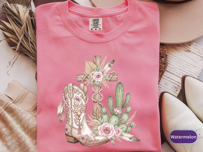 May include: A pink Comfort Colors t-shirt featuring a graphic of a cross, a cowboy boot, and a cactus. The boot is cream-colored with floral and beaded details. The cross is made of wood and adorned with flowers and feathers. The shirt has a crew neck.