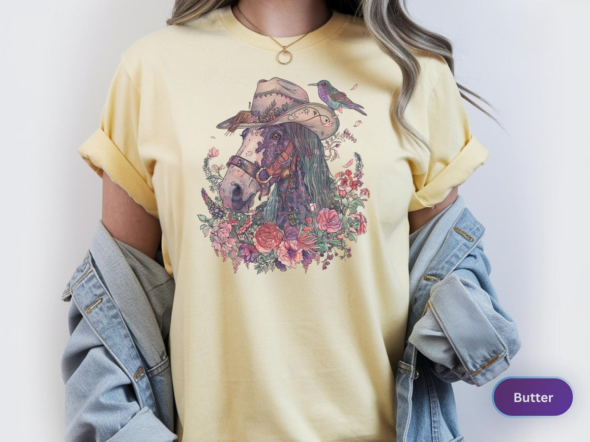 May include: Butter-colored t-shirt featuring a detailed illustration of a horse wearing a cowboy hat, surrounded by flowers and a small bird. The design incorporates shades of purple, pink, and green, creating a whimsical and artistic aesthetic.