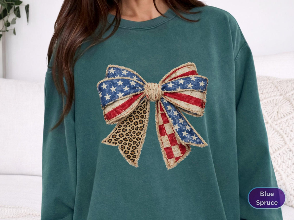 May include: A teal-colored sweatshirt featuring a large bow design. The bow incorporates elements of the American flag with red and white stripes, blue stars, and a leopard print pattern. The sweatshirt has a crew neck and long sleeves.