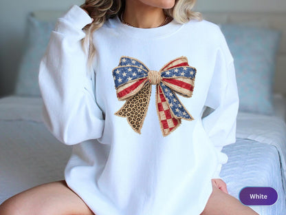 May include: White sweatshirt featuring a large bow design. The bow incorporates elements of the American flag with stars, stripes, and a leopard print accent. The design is centered on the front of the sweatshirt.