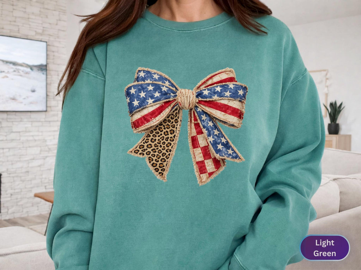 May include: A light green sweatshirt featuring a large bow design. The bow incorporates elements of the American flag with red and white stripes, blue stars, and a leopard print pattern. The sweatshirt is a solid light green color.