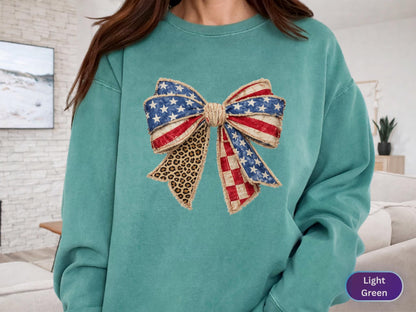 May include: A light green sweatshirt featuring a large bow design. The bow incorporates elements of the American flag with red and white stripes, blue stars, and a leopard print pattern. The sweatshirt is a solid light green color.