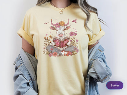 May include: Butter-colored t-shirt featuring a whimsical illustration of a cow reading a book. The cow is adorned with flowers and surrounded by floral elements. The design is in soft pastel colors, creating a charming and artistic aesthetic.