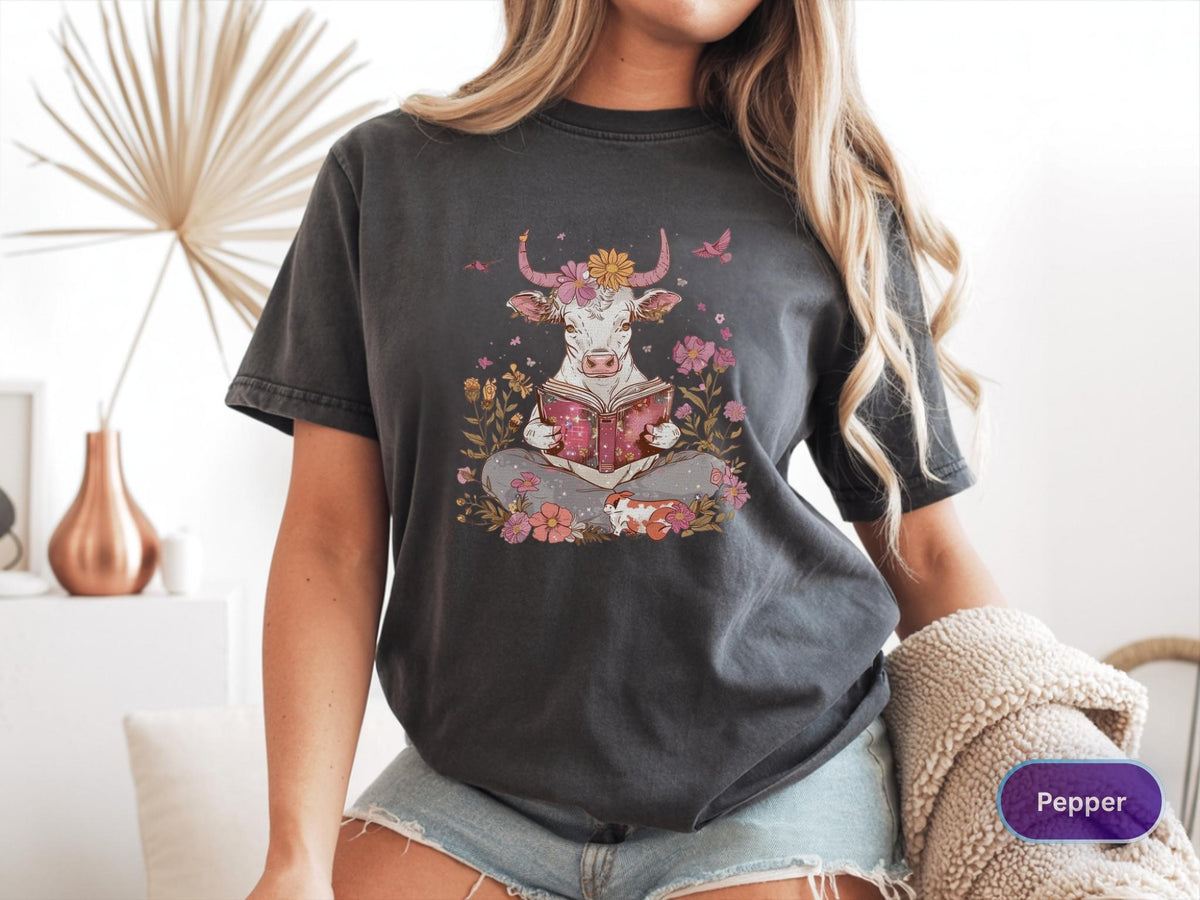 May include: Dark grey t-shirt featuring a whimsical illustration of a cow reading a book, surrounded by flowers and butterflies. The cow has flower horns and is seated in a meditative pose. The shirt is a relaxed fit.
