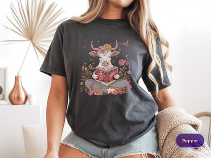 May include: Dark grey t-shirt featuring a whimsical illustration of a cow reading a book, surrounded by flowers and butterflies. The cow has flower horns and is seated in a meditative pose. The shirt is a relaxed fit.