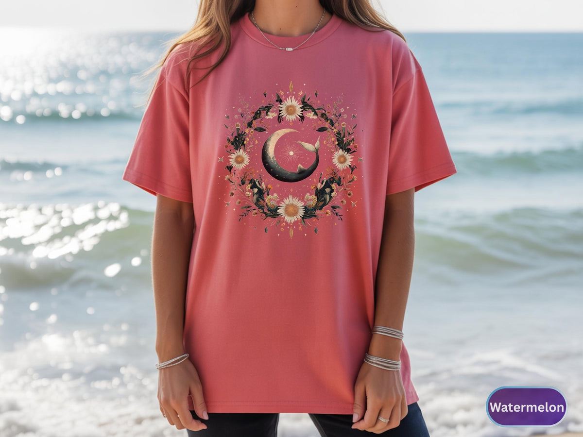 May include: A watermelon-colored t-shirt featuring a floral wreath design with a crescent moon and a whale. The wreath includes sunflowers and other flowers. The shirt is a short-sleeved, crew neck style.