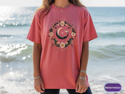 May include: A watermelon-colored t-shirt featuring a floral wreath design with a crescent moon and a whale. The wreath includes sunflowers and other flowers. The shirt is a short-sleeved, crew neck style.