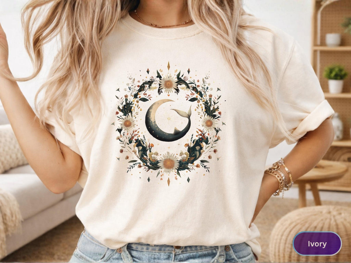 May include: Ivory-colored t-shirt featuring a circular floral design with a crescent moon and whale tail. The design includes white flowers, dark green leaves, and small brown accents. The shirt is a relaxed fit.