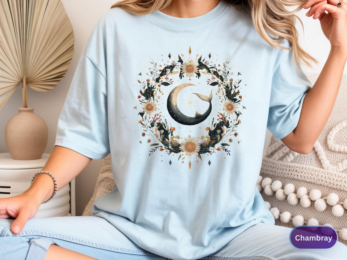 May include: Light blue t-shirt featuring a watercolor design of a crescent moon with a whale tail, surrounded by a floral wreath. The design includes sunflowers and other botanical elements. The shirt is a soft, comfortable material.