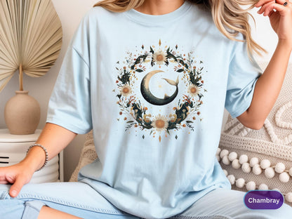 May include: Light blue t-shirt featuring a watercolor design of a crescent moon with a whale tail, surrounded by a floral wreath. The design includes sunflowers and other botanical elements. The shirt is a soft, comfortable material.