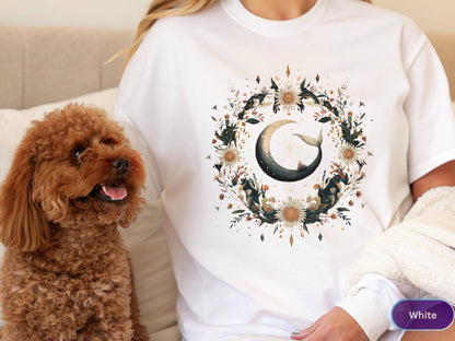 May include: White t-shirt featuring a watercolor design of a crescent moon with a whale tail, surrounded by a floral wreath. The design includes shades of beige, green, and gold. A brown dog is in the foreground.