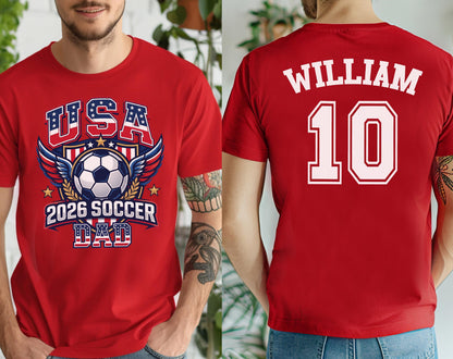 May include: Red t-shirt with a soccer-themed graphic. The front features "USA" in a patriotic design, a soccer ball, wings, and "2026 SOCCER DAD." The back displays "WILLIAM" above the number "10" in white.