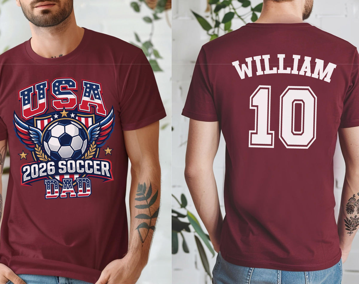 May include: Maroon t-shirt with a soccer-themed graphic. The front features "USA" in red, white, and blue, a soccer ball, wings, and "2026 SOCCER DAD." The back displays "WILLIAM" and the number "10" in white.