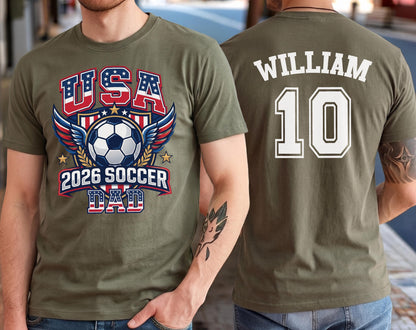 May include: Olive green t-shirt with a soccer-themed graphic. The front features "USA" in red and blue, a soccer ball, wings, and "2026 SOCCER DAD." The back displays "WILLIAM" and the number "10" in white.