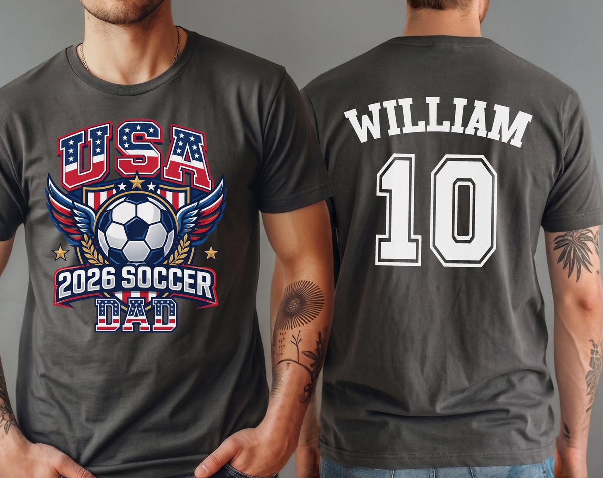 May include: Dark gray t-shirt with a soccer-themed graphic. The front features "USA" in red and blue, a soccer ball, wings, and "2026 SOCCER DAD." The back displays "WILLIAM" and the number "10" in white.