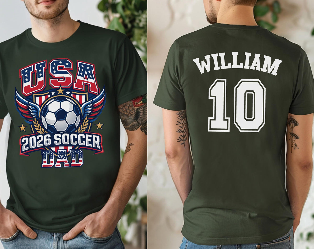 May include: Dark green t-shirt with a soccer-themed graphic. The front features "USA" in red, white, and blue, a soccer ball, wings, and "2026 SOCCER DAD." The back displays "WILLIAM" and the number "10" in white.