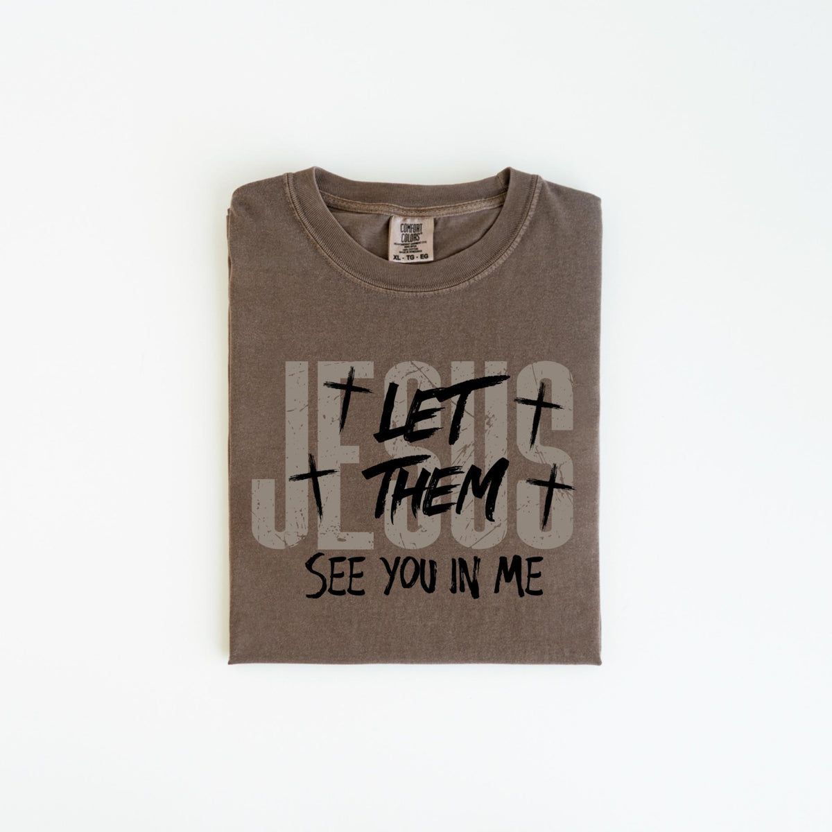 May include: A folded, brown t-shirt with the text "JESUS LET THEM SEE YOU IN ME" printed in a distressed, gray and black font. The shirt has a crew neck and a tag at the neckline.