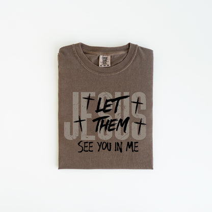 May include: A folded, brown t-shirt with the text "JESUS LET THEM SEE YOU IN ME" printed in a distressed, gray and black font. The shirt has a crew neck and a tag at the neckline.