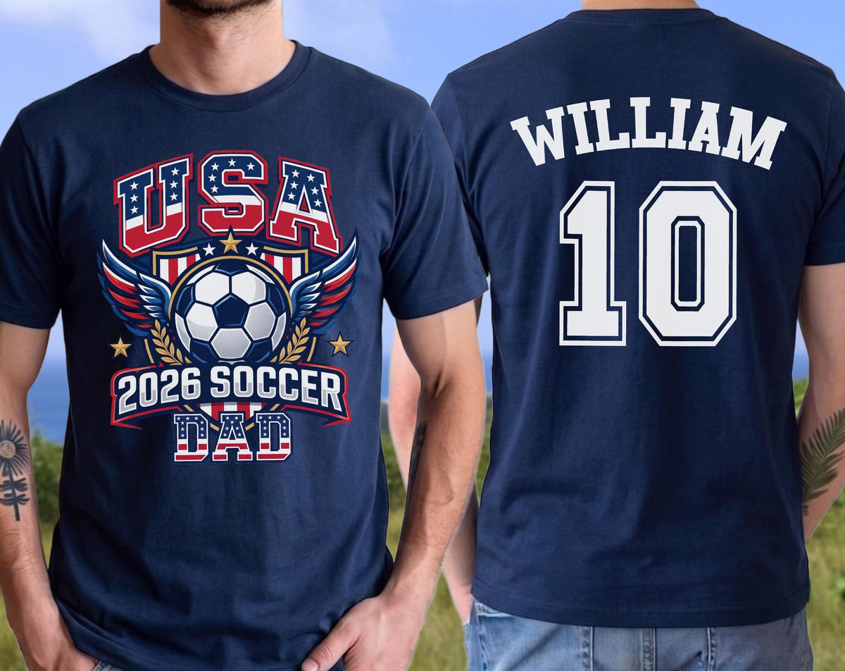 May include: Navy blue t-shirt with a soccer-themed graphic. The front features "USA" in red and blue, a soccer ball, wings, and "2026 SOCCER DAD." The back displays "WILLIAM" and the number "10" in white.