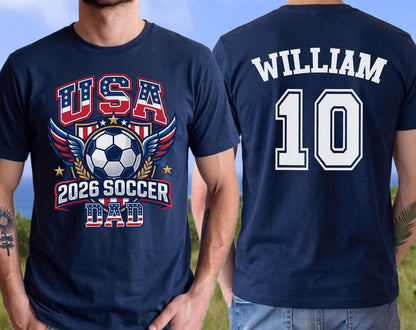 May include: Navy blue t-shirt with a soccer-themed graphic. The front features "USA" in red and blue, a soccer ball, wings, and "2026 SOCCER DAD." The back displays "WILLIAM" and the number "10" in white.