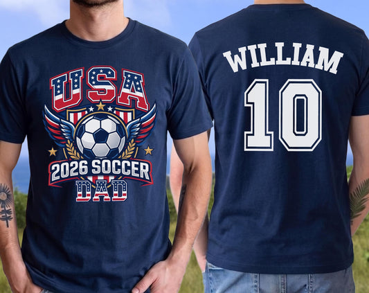 May include: Navy blue t-shirt with a soccer-themed graphic. The front features "USA" in red and blue, a soccer ball, wings, and "2026 SOCCER DAD." The back displays "WILLIAM" and the number "10" in white.