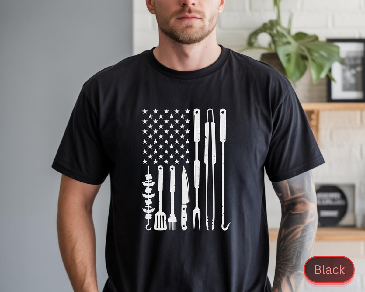 May include: Black t-shirt featuring a white graphic of the American flag with grilling tools replacing the stripes. The design includes a skewer, spatula, knife, fork, and tongs. The shirt is a crew neck.