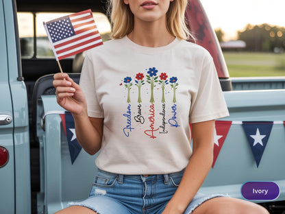 May include: Ivory-colored t-shirt featuring a floral design with red and blue flowers and the words "Freedom, Brave, America, Independence, Power." An American flag is held in the hand. The shirt is paired with blue denim shorts.