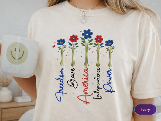 May include: Ivory-colored t-shirt featuring a floral design with red and blue flowers. The words "Freedom, Brave, America, Independence, Power" are vertically aligned. A white mug with a smiley face is held in the left hand.