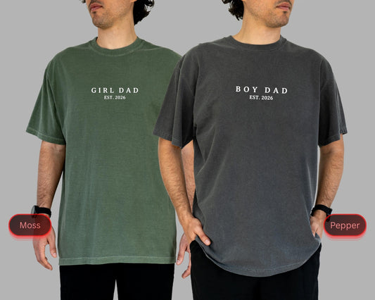 May include: Two short-sleeved t-shirts, one moss green and one pepper gray. The moss green shirt has the text "GIRL DAD EST. 2026" printed in white. The pepper gray shirt has the text "BOY DAD EST. 2026" printed in white.