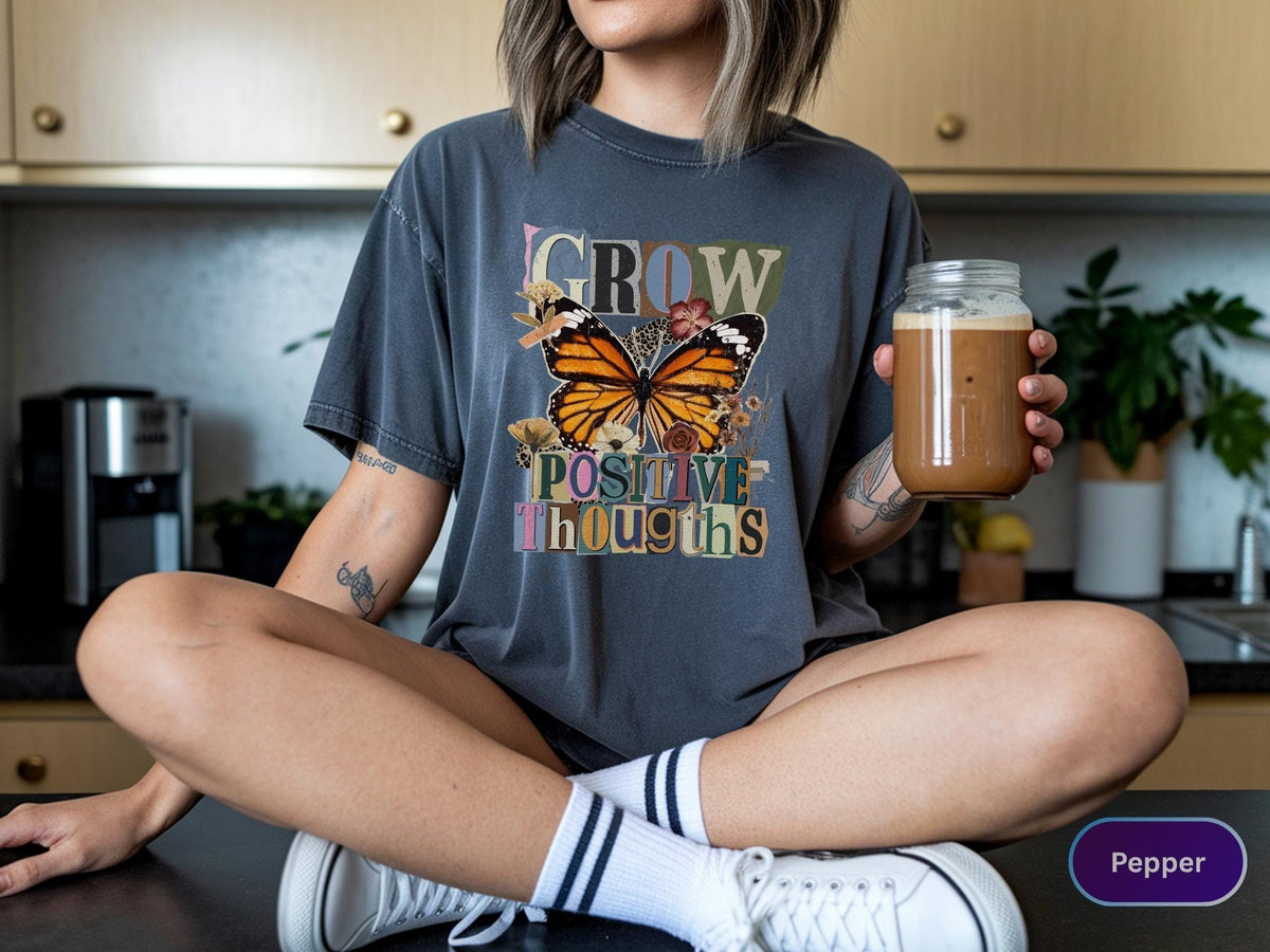 May include: Dark gray t-shirt with the words "GROW POSITIVE THOUGHTS" and a butterfly graphic. The butterfly has orange, black, and white wings. The person is wearing white sneakers and striped socks. A glass jar with a brown liquid is held in the hand.