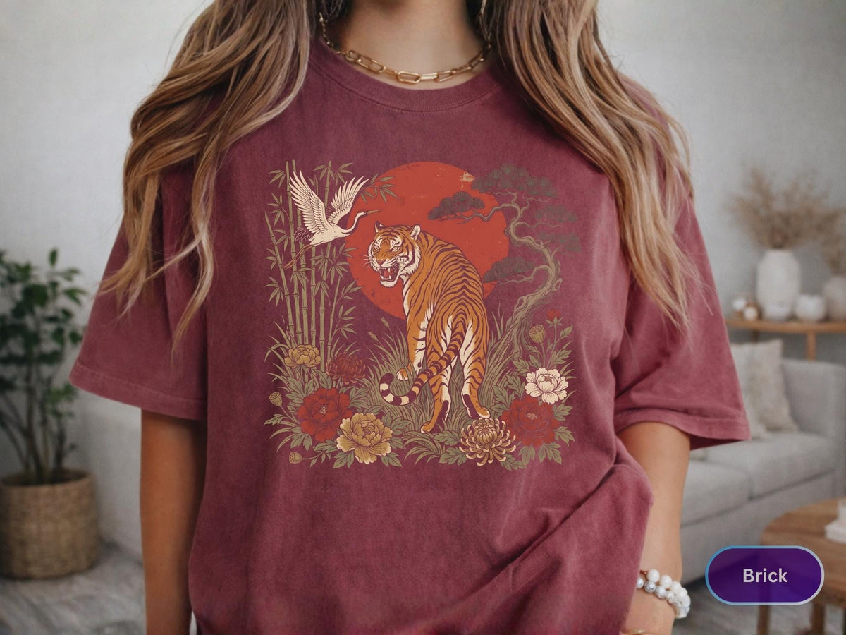 May include: Brick-colored t-shirt featuring a detailed illustration of a tiger, crane, and floral elements set against a red sun. The design has an Asian-inspired aesthetic. The shirt is a short-sleeved crew neck.