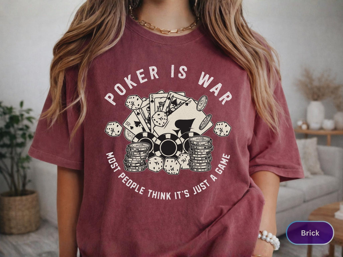 May include: Brick-colored t-shirt with the text "POKER IS WAR" above a graphic of playing cards, dice, and poker chips. The text "MOST PEOPLE THINK IT'S JUST A GAME" is below the graphic. The shirt has short sleeves.