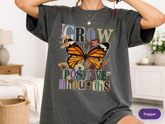 May include: Dark gray t-shirt featuring a large butterfly with orange and black wings, surrounded by flowers. The shirt has the words "GROW POSITIVE THOUGHTS" in a collage-style design. The shirt has short sleeves.