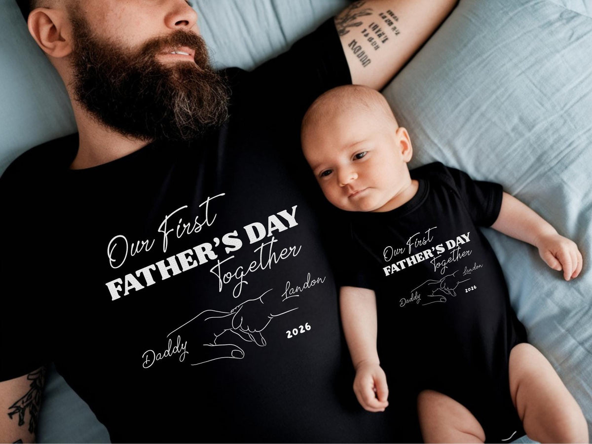 May include: Black matching t-shirts and onesie with the text "Our First Father's Day Together" in white script. The design includes a line drawing of two hands touching. The onesie is for a baby.