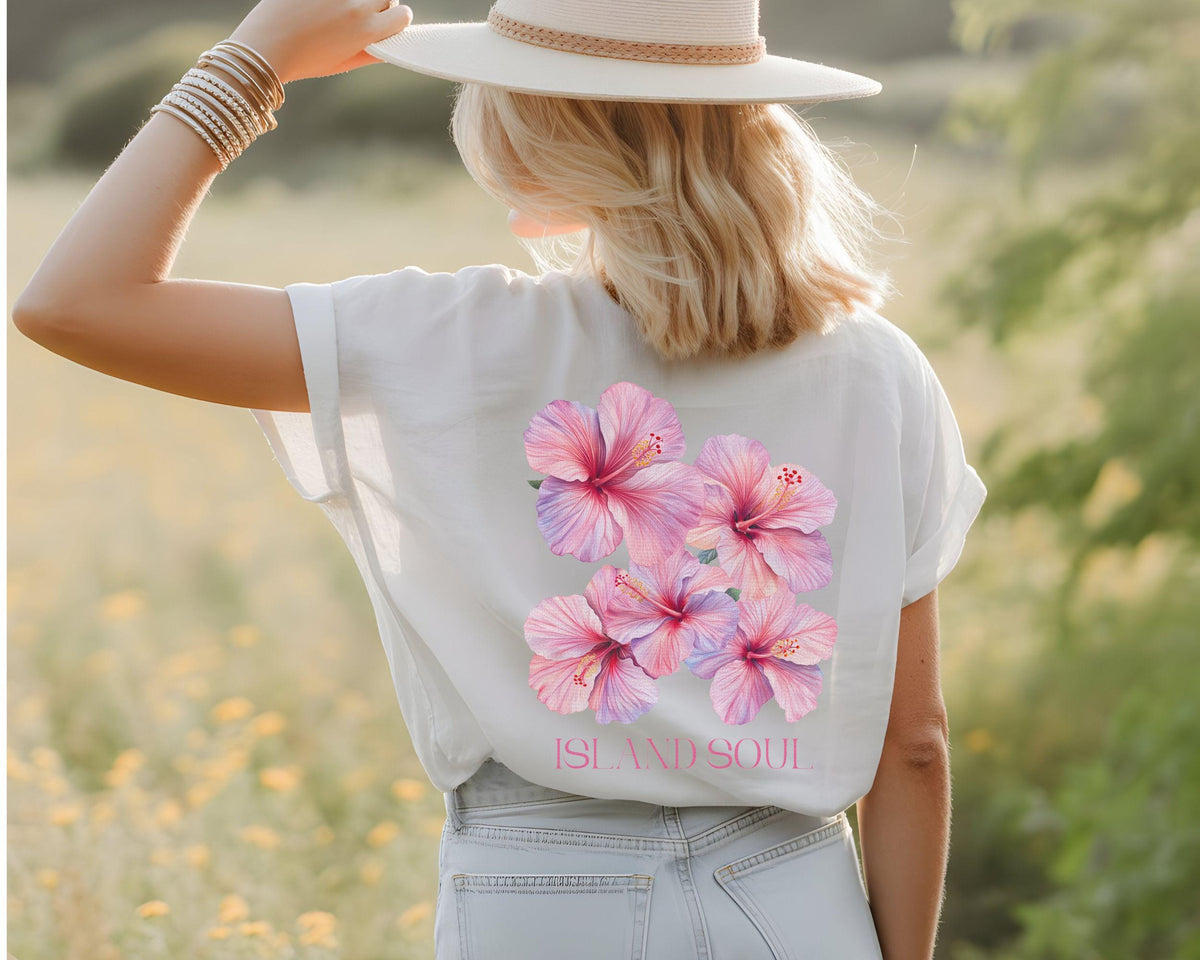 May include: White t-shirt featuring a cluster of pink hibiscus flowers with red centers. The text "ISLAND SOUL" is printed below the flowers. The person is wearing a white hat and blue jeans.