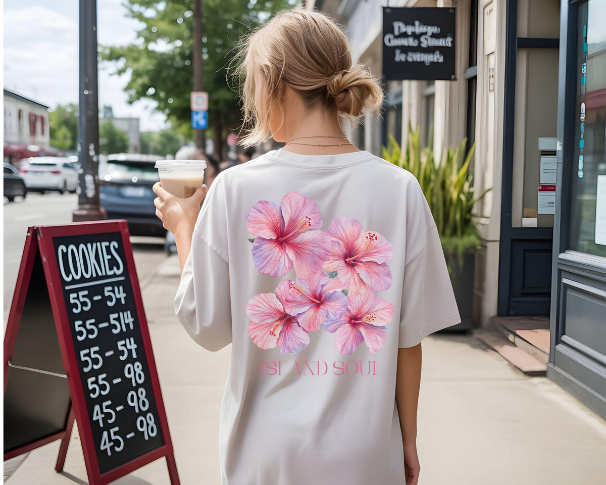 May include: A white t-shirt featuring a cluster of pink hibiscus flowers with purple accents and the text "ISLAND SOUL". The shirt is worn outdoors, with a sign for cookies in the background.